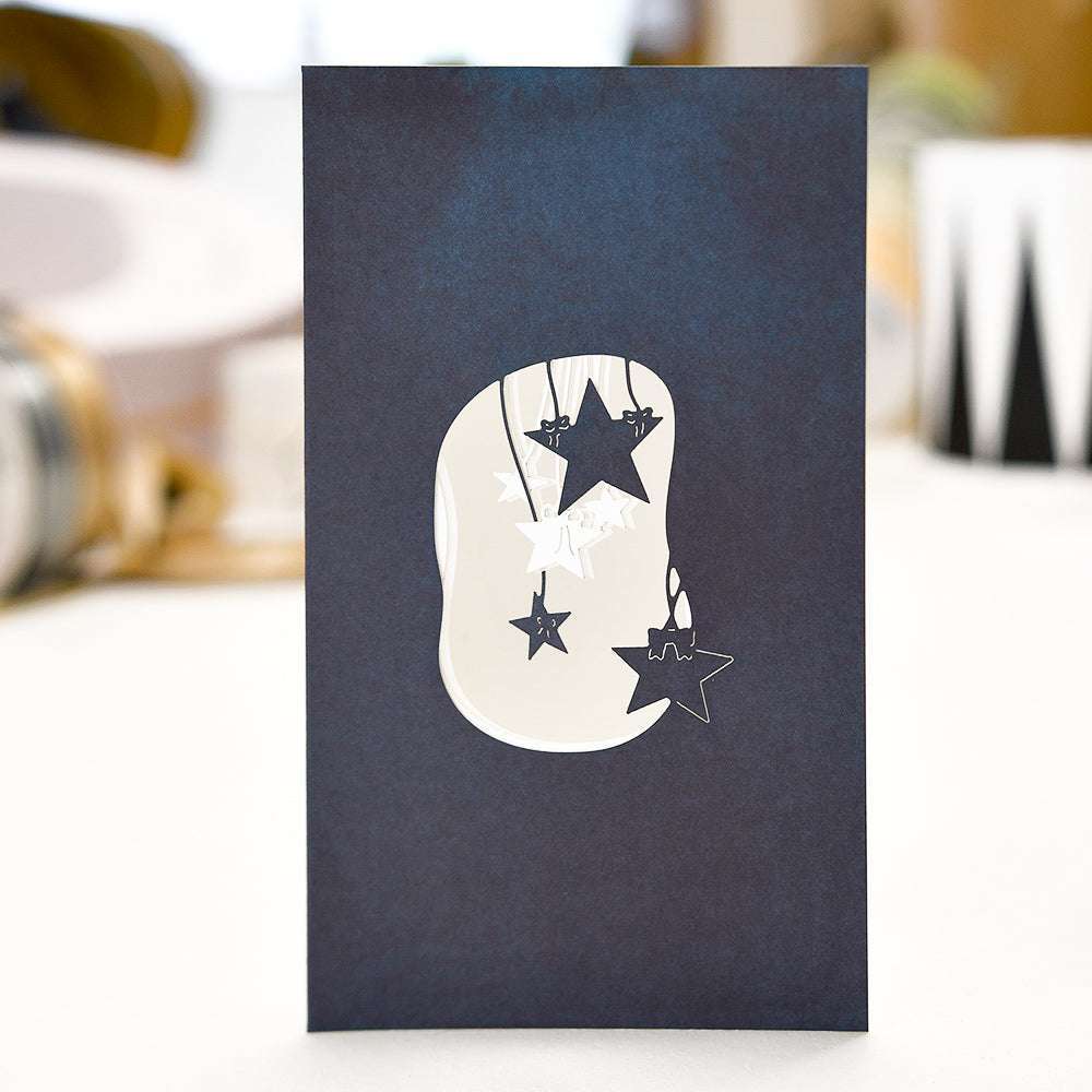 Eine marineblaue Karte von Alexandra Renke mit an Schnüren hängenden sternförmigen Ausschnitten, die aufrecht auf einer hellen Fläche stehen.