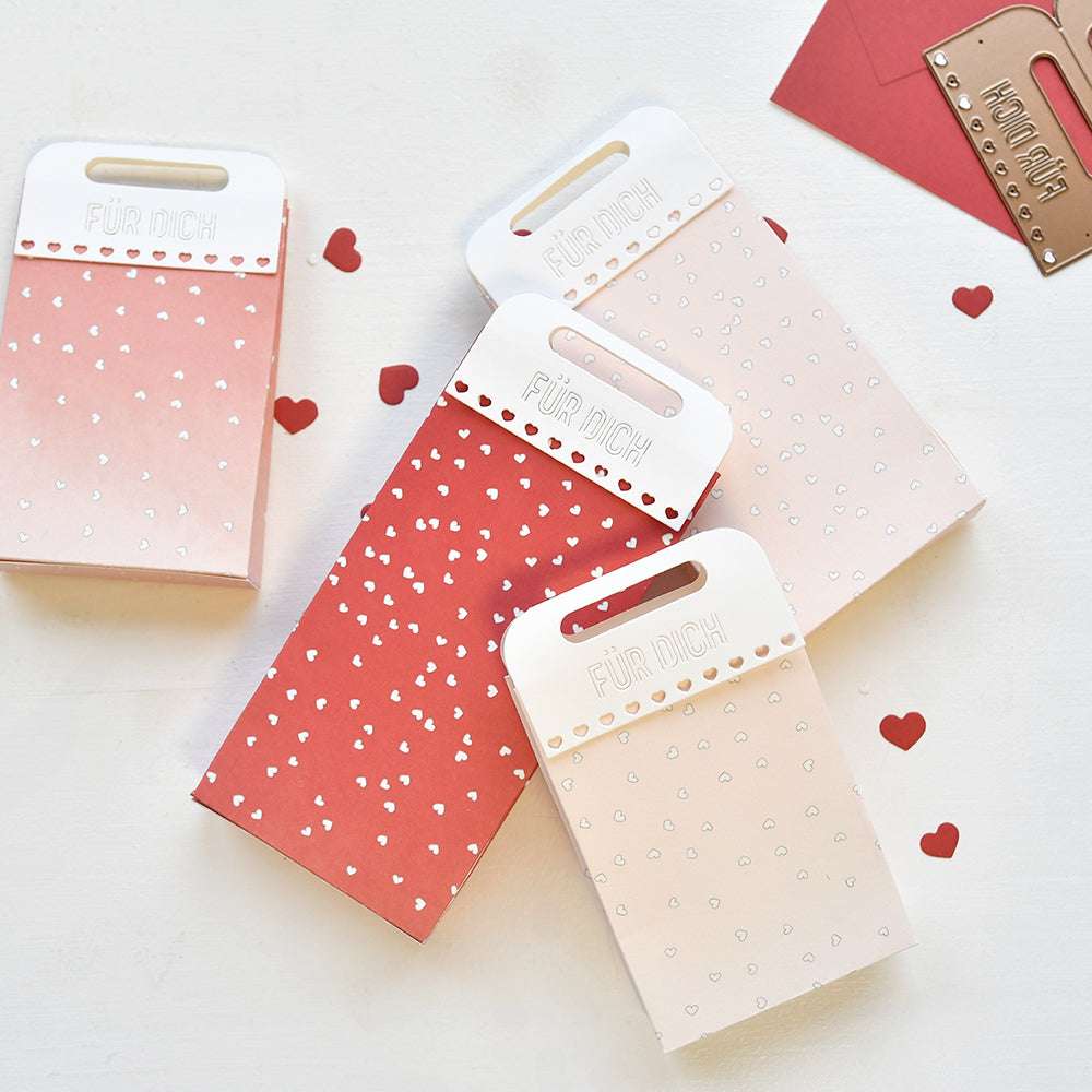 Vier rosafarbene und rote Alexandra Renke Geschenktüten mit weißen Herzmustern und "Für Dich"-Etiketten, umgeben von Papierherzen - eine auffällige, handgefertigte Verpackung, perfekt zum Verschenken von Liebe.