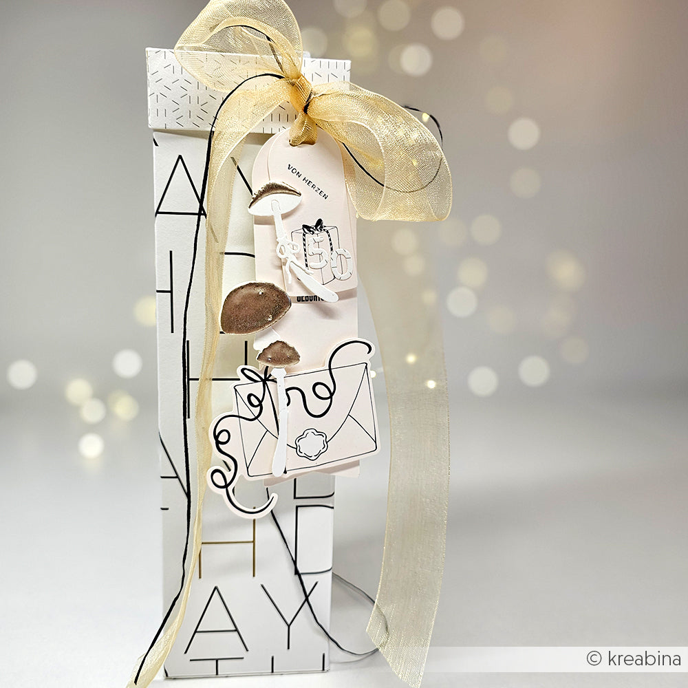 Ein großes Geschenk, verpackt in weißem Papier mit fetten Buchstaben, goldenem Band und dekorativen Anhängern mit Illustrationen - perfekt für alle, die Alexandra Renke oder einzigartige Kollektionen lieben.