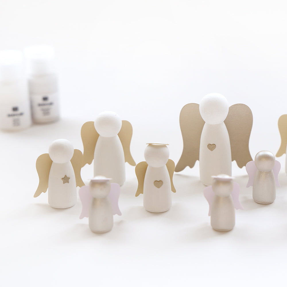 Alexandra Renke Holzfiguren in Form von Engeln, verziert mit aufgemalten Flügeln, Herzen und Sternen, bilden eine bezaubernde Weihnachtsdekoration auf einer weißen Fläche mit Flaschen im Hintergrund.