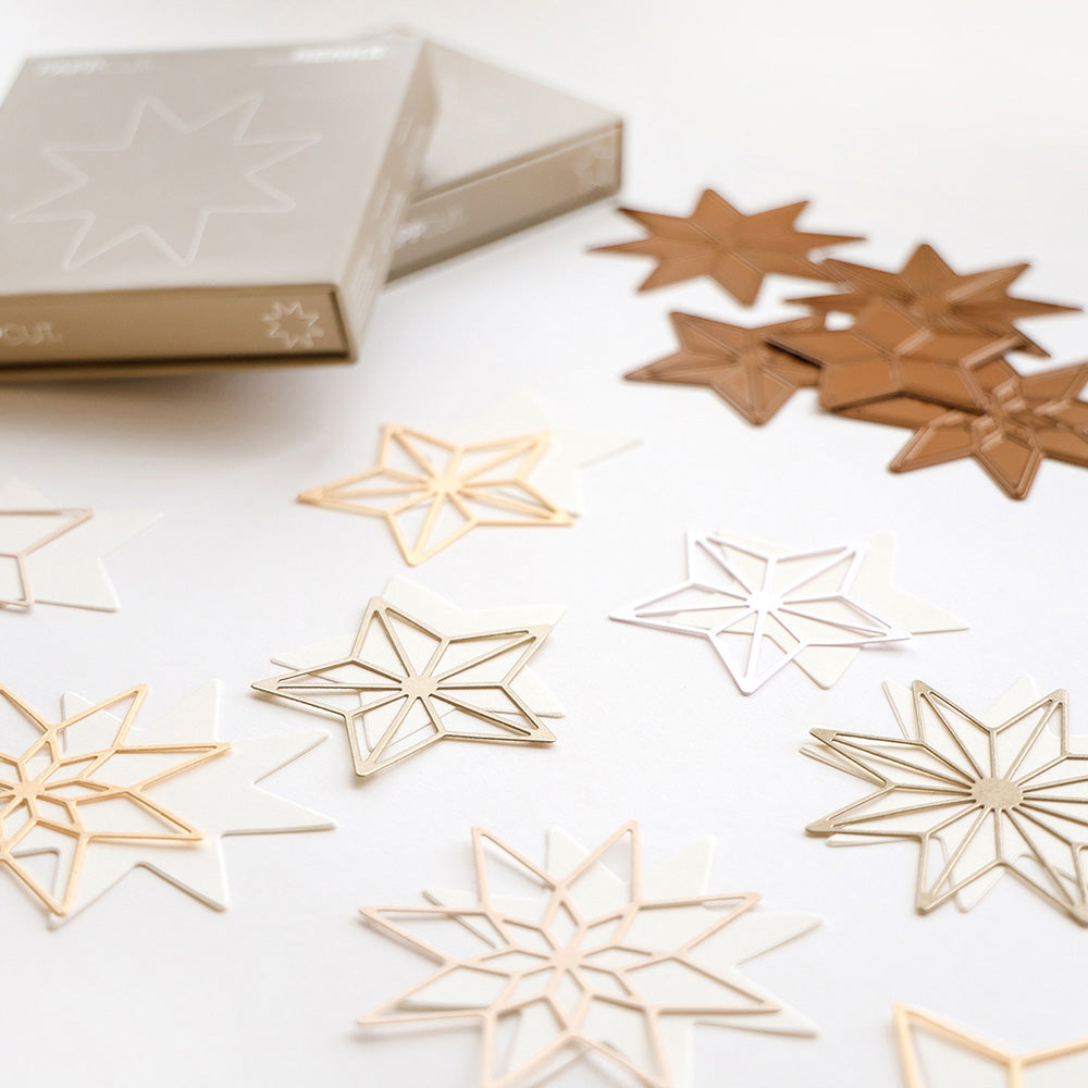 Metallische und weiße sternförmige Ornamente von Alexandra Renke sind auf einer weißen Fläche neben zwei beigen Schachteln verstreut und inspirieren zu festlichen Kollektionen.