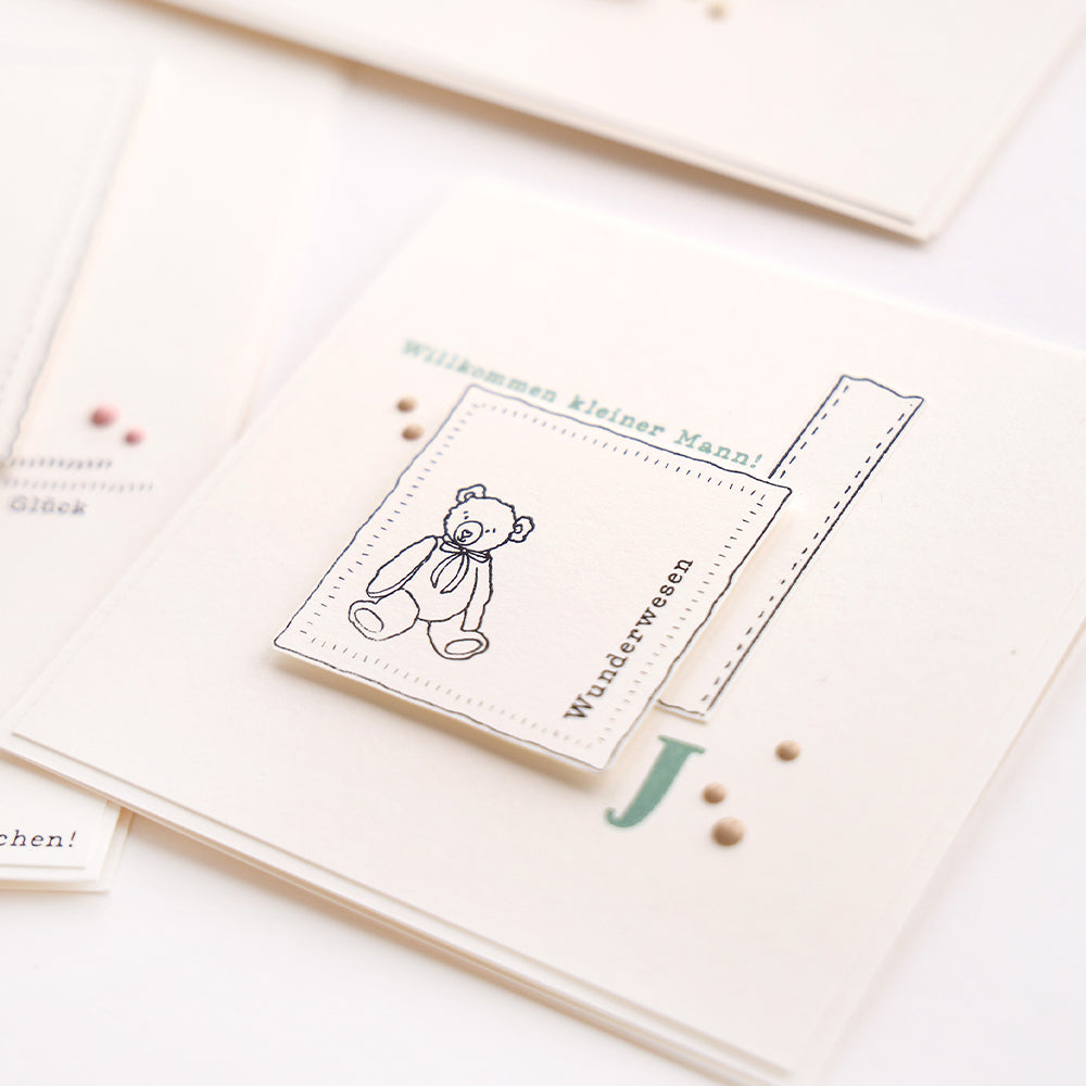 Eine weiße Karte von Alexandra Renke mit einem Teddybär-Motiv, einem Lineal und einem deutschen Begrüßungstext für einen kleinen Jungen - ideal für Kollektionen, die Neuanfänge und Kreativität feiern.