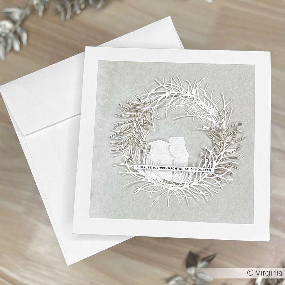Eine weiße Urlaubskarte von Alexandra Renke mit einem silbernen Kranz und zwei Häusern wird mit einem passenden Umschlag geliefert. Dieses charmante Umschläge und Karten Set wird auf einer hellen Oberfläche präsentiert.
