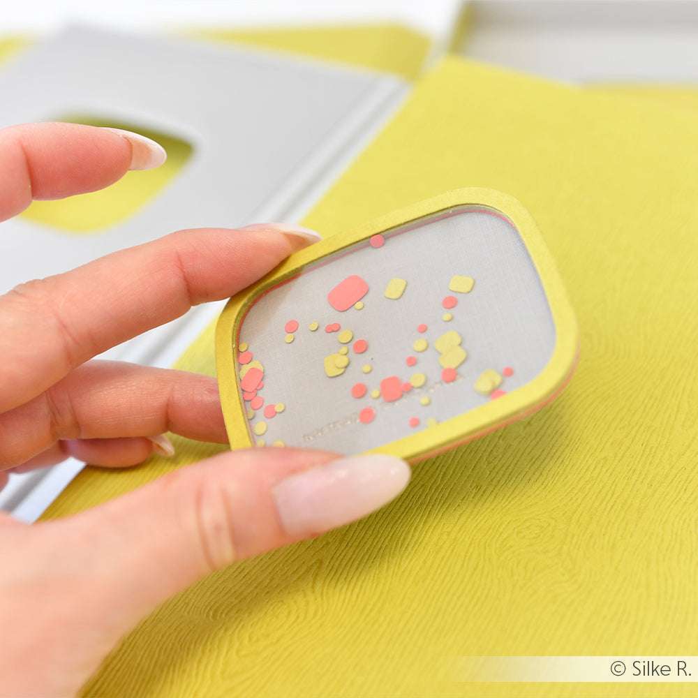 Die Hand hält eine kleine, quadratische Alexandra Renke Leinwand mit buntem Konfetti vor einem gelben, strukturierten Hintergrund - ideal, um Ihre Kreativität oder neue Kollektionen zu präsentieren.