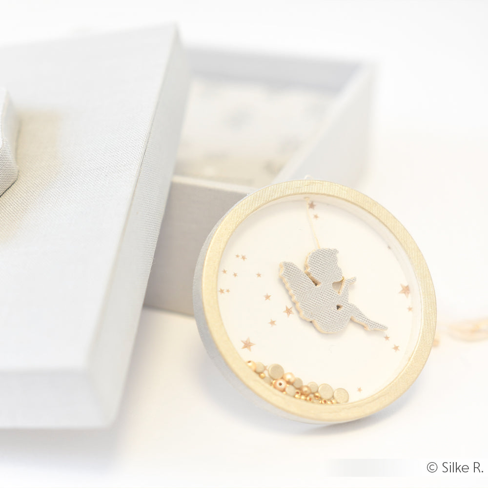 Ein rundes Ornament von Alexandra Renke mit einer Engelssilhouette und goldenen Perlen, das in einer hellgrauen Geschenkbox präsentiert wird.
