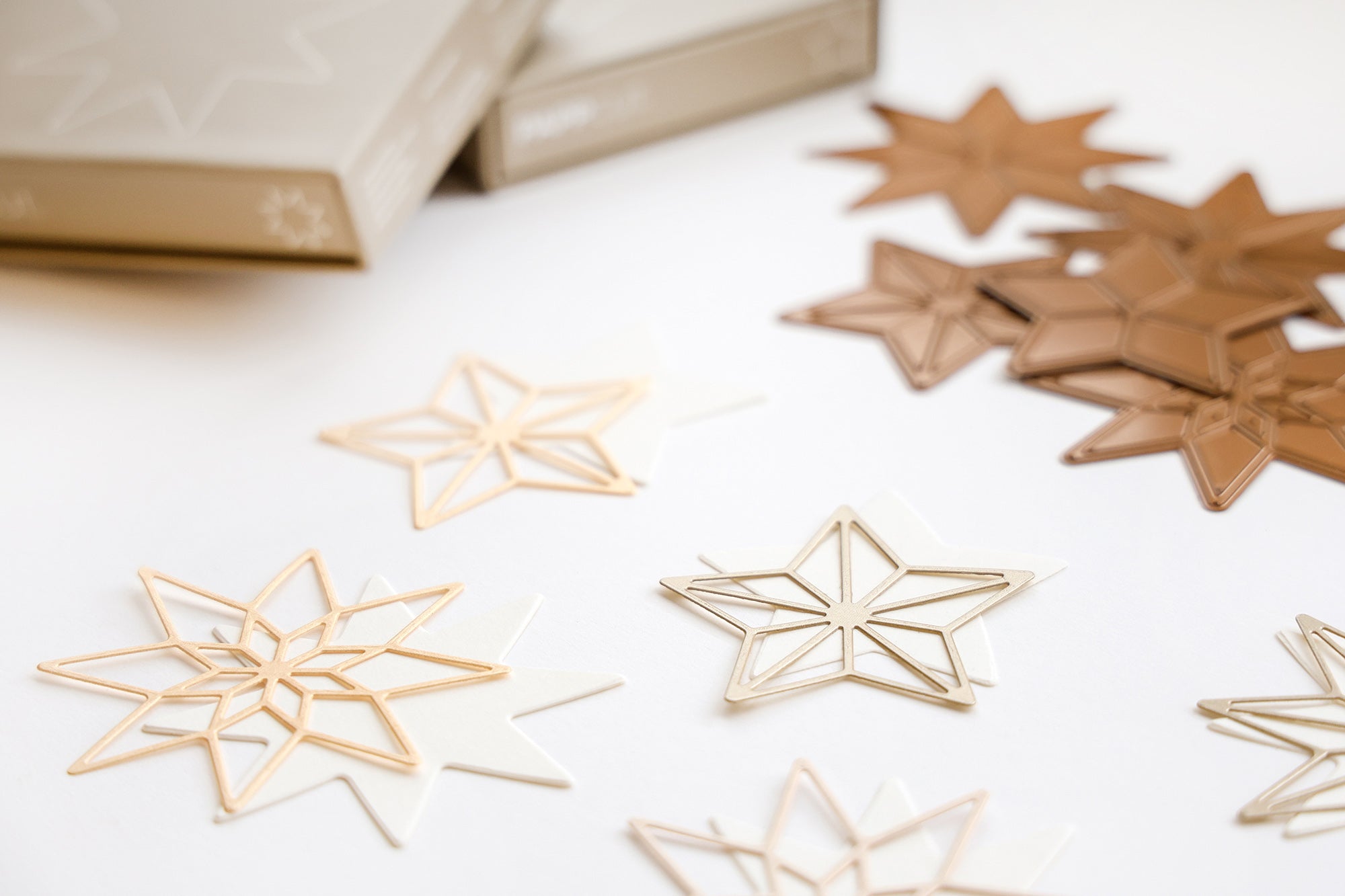 Goldene, bronzene und weiße sternförmige Ornamente sind auf einer weißen Fläche in der Nähe von beigen Kästen verstreut.