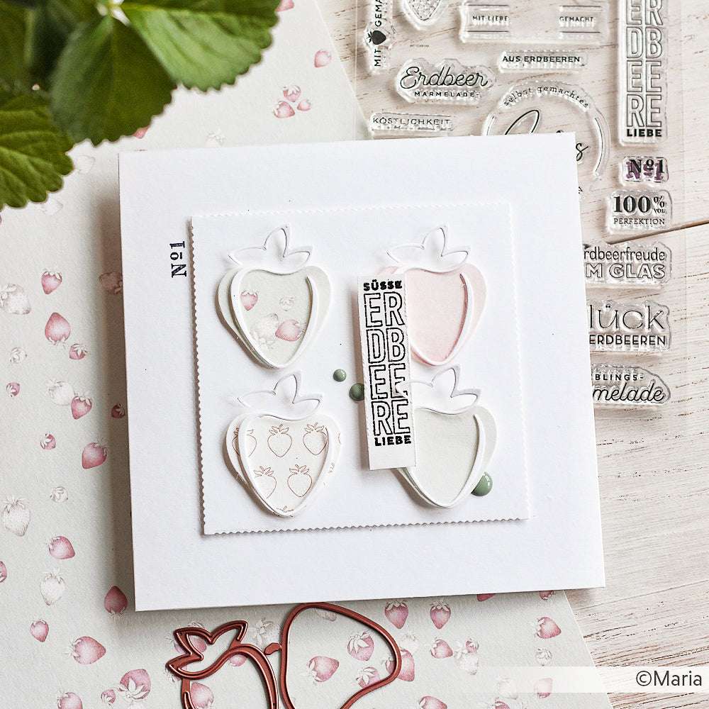 Eine handgefertigte Karte mit Erdbeermotiven, die mit der Alexandra Renke Motivstanze Erdbeere, Stempeln und Verzierungen auf einer weißen Holzoberfläche hergestellt wurde.