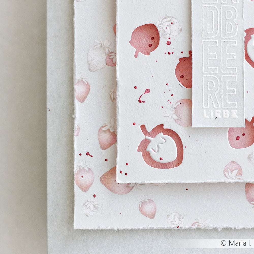 Lagenweise weiße Designpapiere von Alexandra Renke zeigen rosafarbene Erdbeerprints und die Prägung "ERDBEERE LIEBE", ergänzt durch charmante Details aus der Motivstanze Erdbeer Etikett.