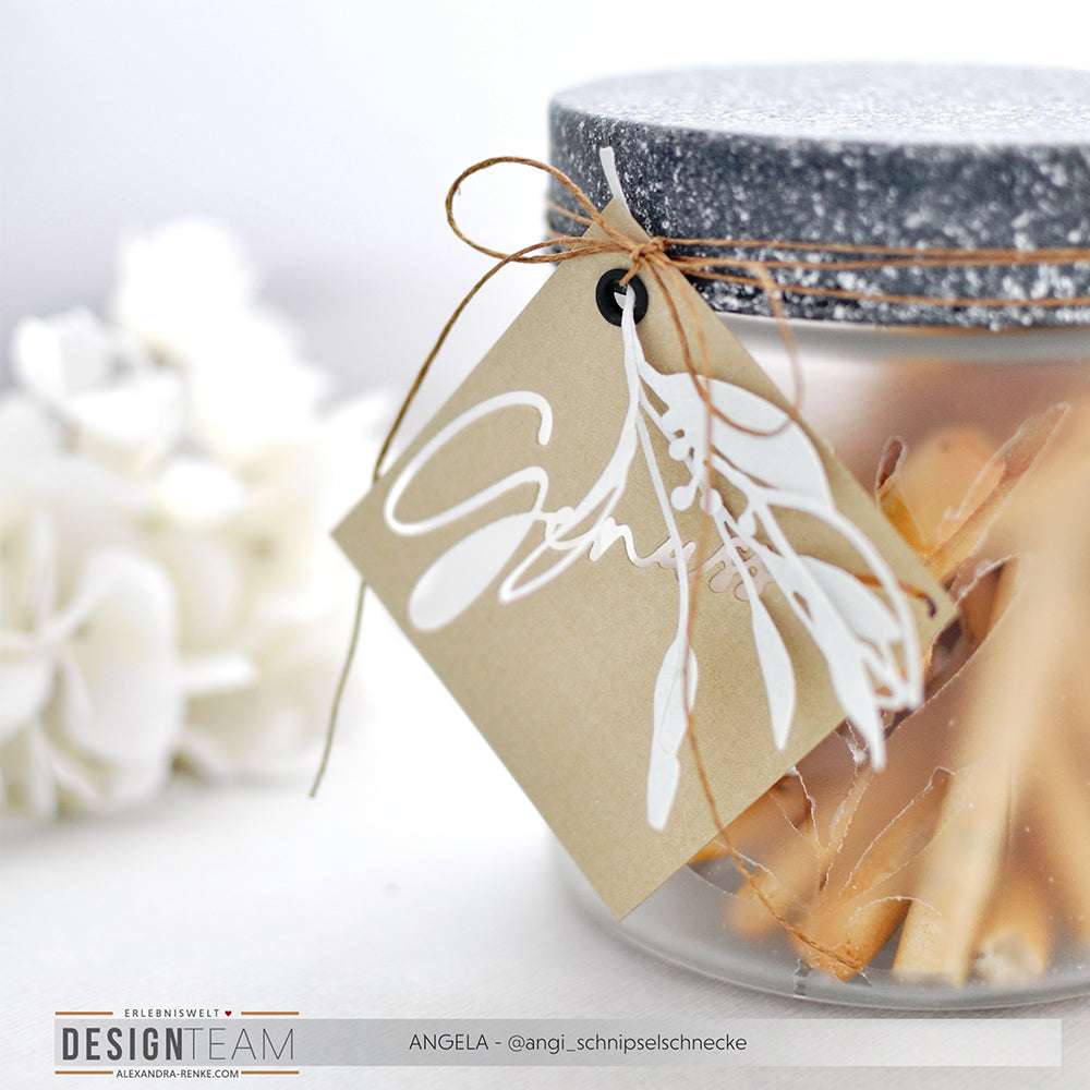 Ein Glas von Alexandra Renke mit Rankenanhänger und weißem Blattmuster ist mit Zimtstangen gefüllt. Weiße Blumen und Designpapiere, die mit der Motivstanze Salbei gestaltet wurden, sorgen im Hintergrund für Charme.