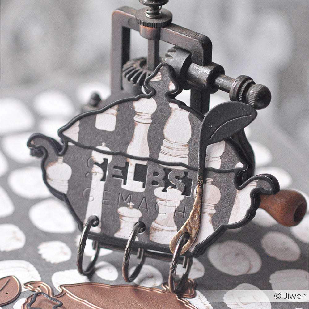 Ein dekorativer Metallclip hält eine Karte im Vintage-Stil aus Alexandra Renke Designpapier mit Schachfigurenmotiven und den Buchstaben SELBST GEMACHT.