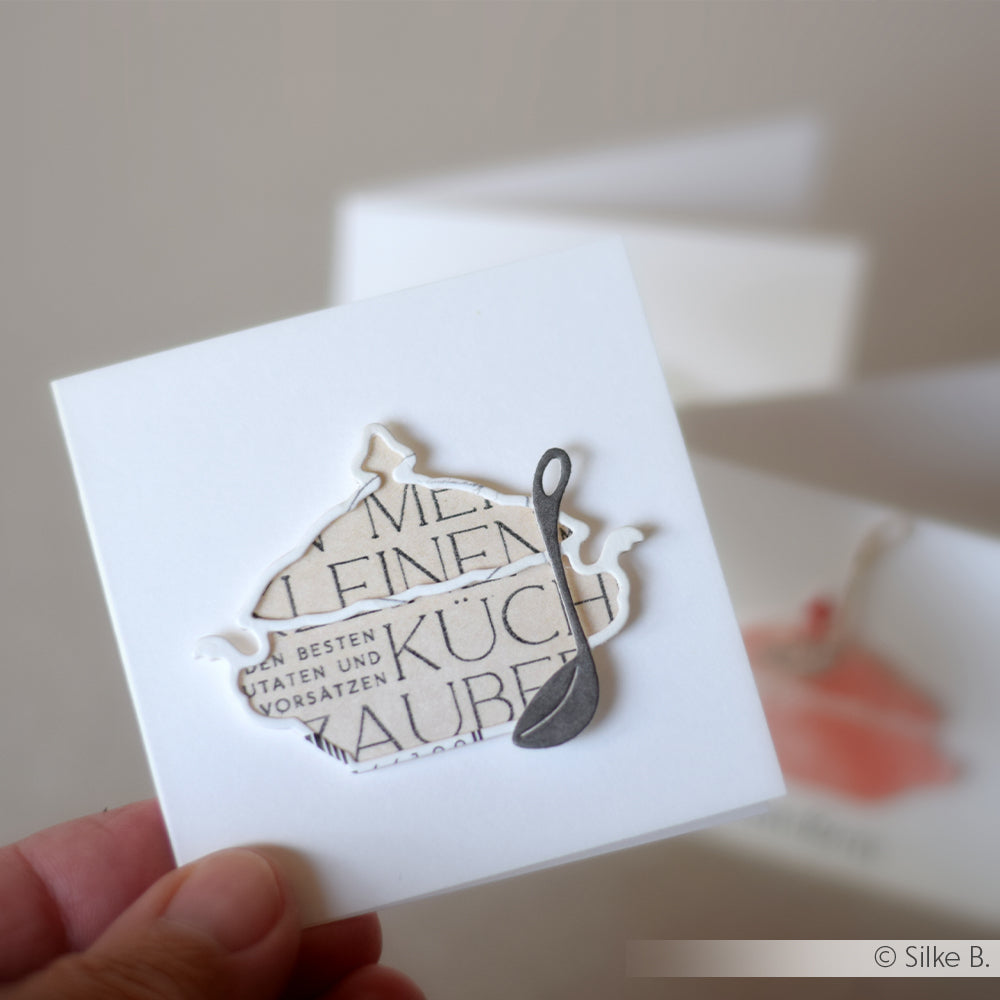 Eine Hand hält eine Karte mit einem Teekannen- und Schöpfkellenmotiv, die mit der Alexandra Renke Motivstanze Terrinen Duo Medium aus weißem Papier gestaltet wurde.