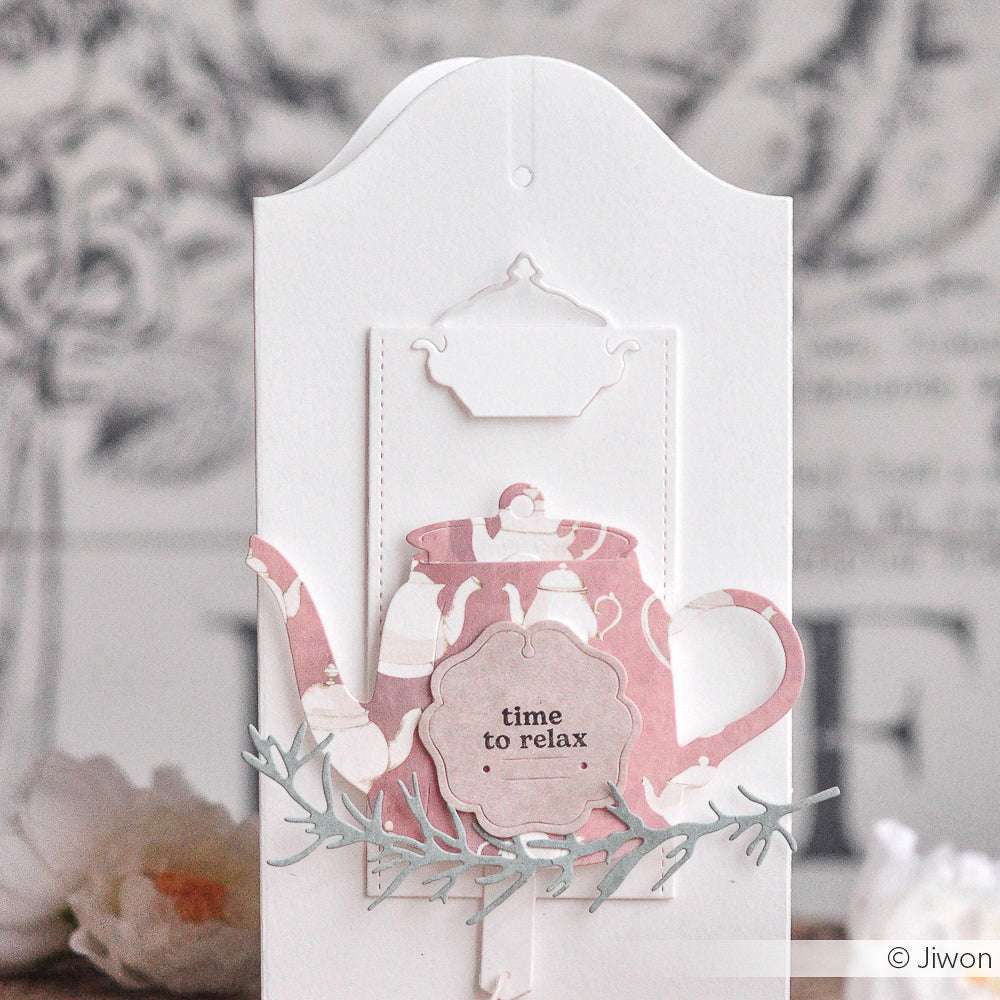 Eine rosa-weiße Teekanne mit der Aufschrift "Zeit zum Entspannen" steht auf einer eleganten weißen Karte mit floralen Akzenten, die aus exquisiten Designpapieren von Alexandra Renke gefertigt wurde.