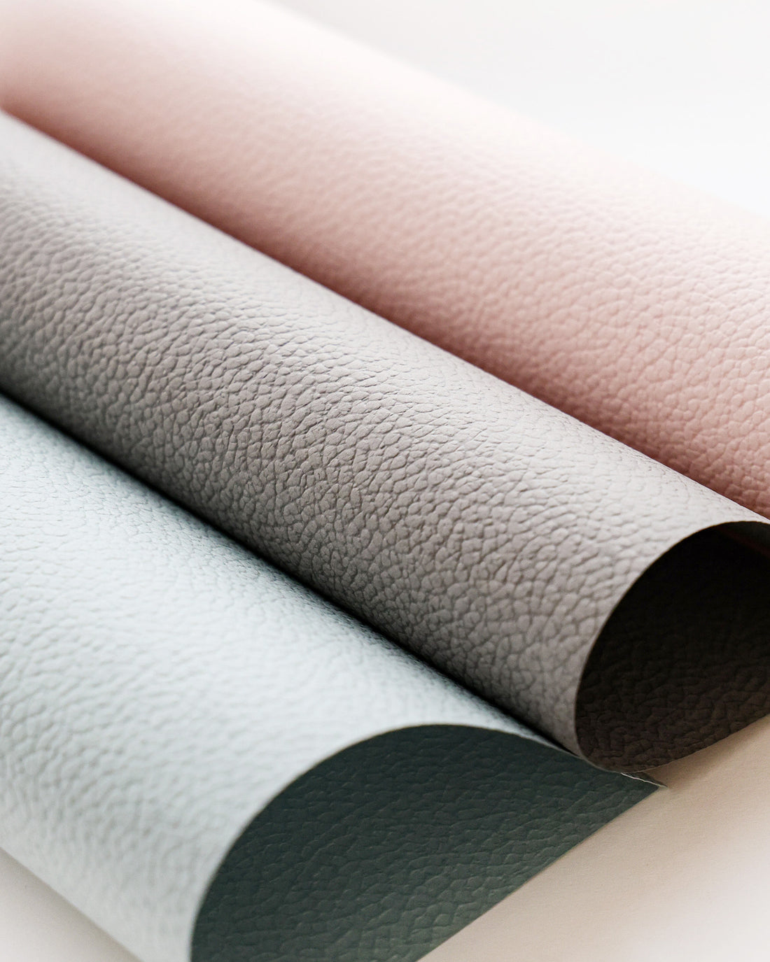 Drei strukturierte Papierbögen in Pastellrosa, Grau und Blau werden aufgerollt und nebeneinander gelegt.