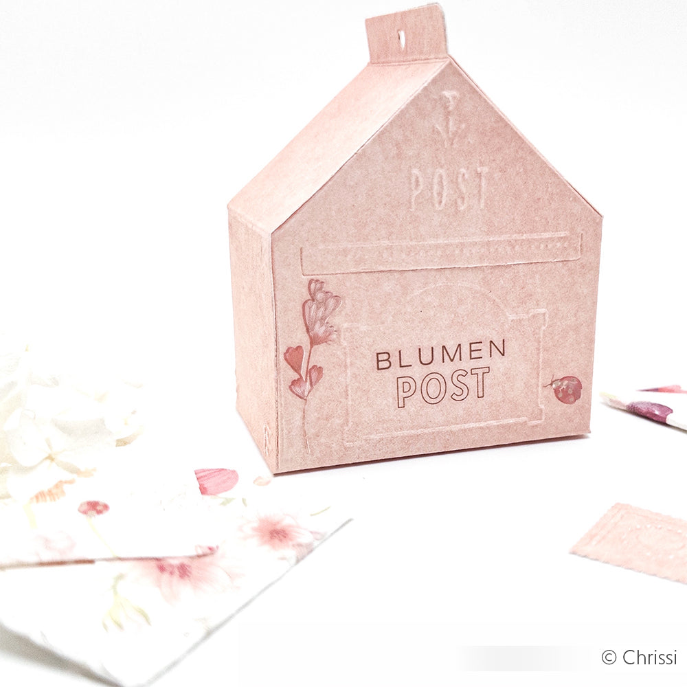 Die Briefkastenbox Blumen Post von Alexandra Renke in Form eines rosa Hauses mit floralen Motiven liegt auf einer weißen Fläche neben Papierstücken - ideal zum Aufbewahren von Stanzformen oder zum Präsentieren Ihrer Cut & Emboss Folder-Kreationen.