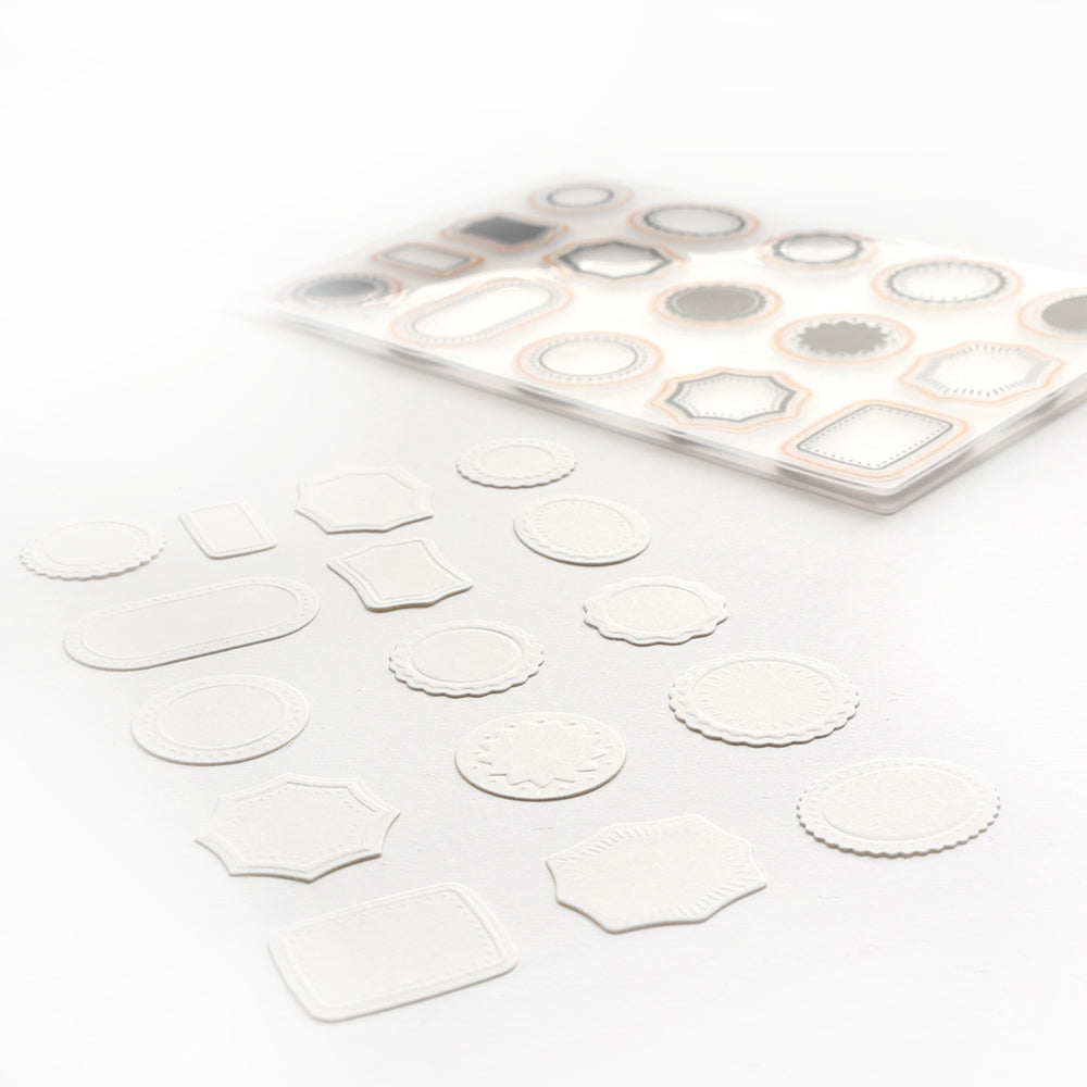 Ein Satz weißer, geprägter Papieretiketten in verschiedenen Formen von Alexandra Renke wird mit einem Bogen passender Klebeetiketten gezeigt, die mit einem Cut & Emboss Folder hergestellt wurden.