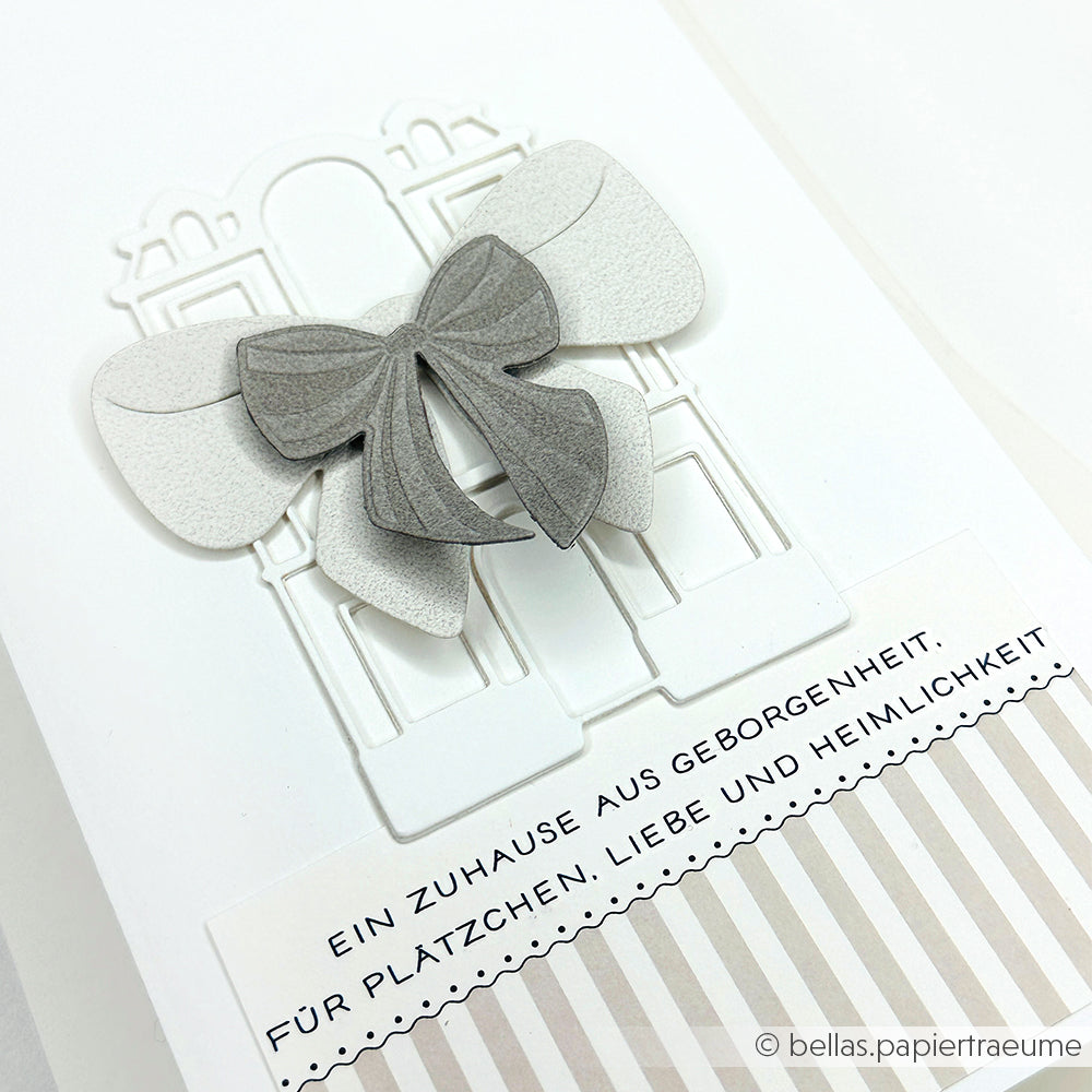 Weiße Karte mit einer grauen 3D-Schleife, die mit Alexandra Renke Schleifenpracht Stanzsysteme hergestellt wurde, einem geprägten Gebäudeumriss und einem deutschen Text über Heimat und Sicherheit - hergestellt mit einem Cut & Emboss Folder für elegante Details.