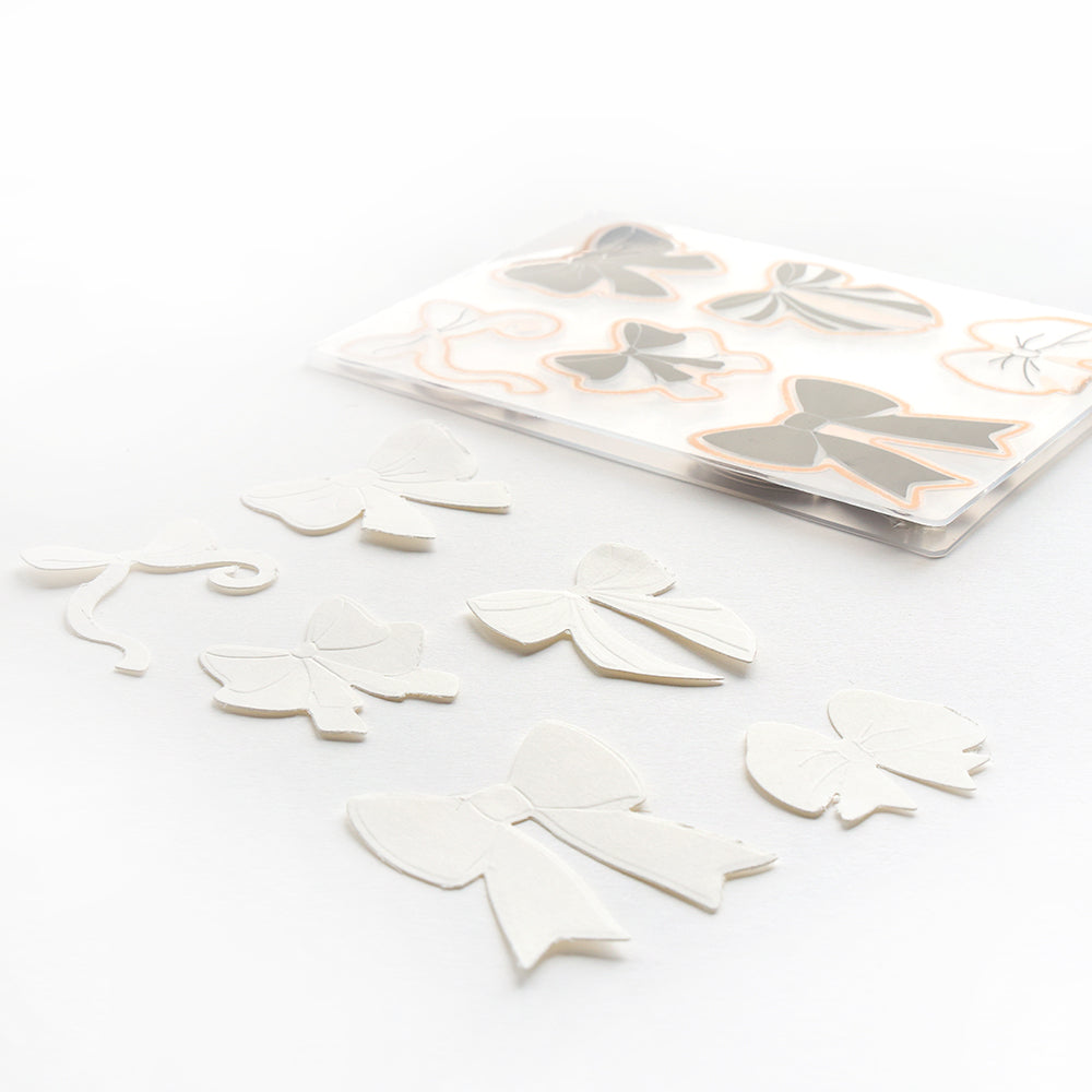 Weiße Papierbögen zum Ausschneiden und ein durchsichtiger Umschlag mit weiteren Bögen auf weißer Oberfläche, alles hergestellt mit dem Alexandra Renke Schleifenpracht Stanzsystem und dem Cut & Emboss Folder für präzise, elegante Designs.