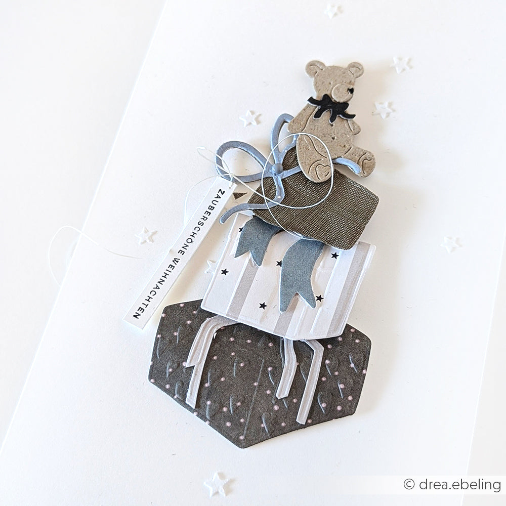 Handgemachte Karte von Alexandra Renke mit geschichteten Papierformen - Teddybär, blaues Band und gemusterte Geschenkschachteln - auf einem strahlend weißen Hintergrund, hergestellt mit einem Cut & Emboss Folder.