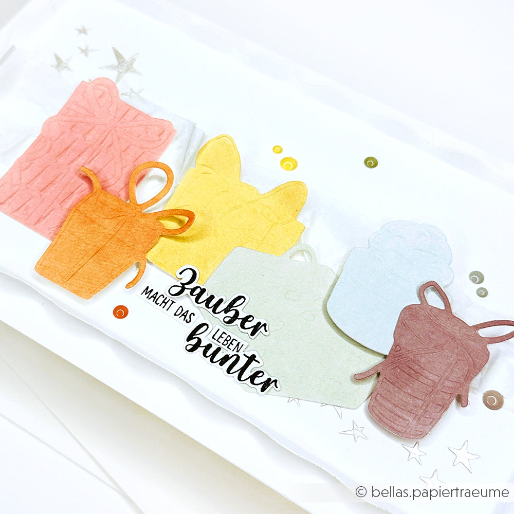 Alexandra Renke ziert mit Stanzsystemen und glitzernden Pailletten eine Karte mit dem Text "Zauber macht das Leben bunter" aus buntem Geschenkpapier.