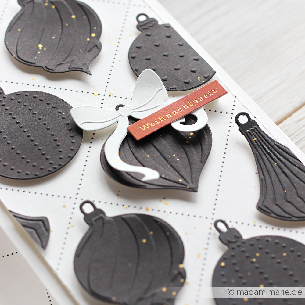 Handgefertigte Karte mit schwarzen Papierornamenten, die mit dem Schneide- und Prägefolder von Alexandra Renke hergestellt wurden, einem Weihnachtszeit-Anhänger und einer weißen Schleife - ideal für die Präsentation von Festtagskugeln mit eleganten Alexandra Renke Stanzsystemen.