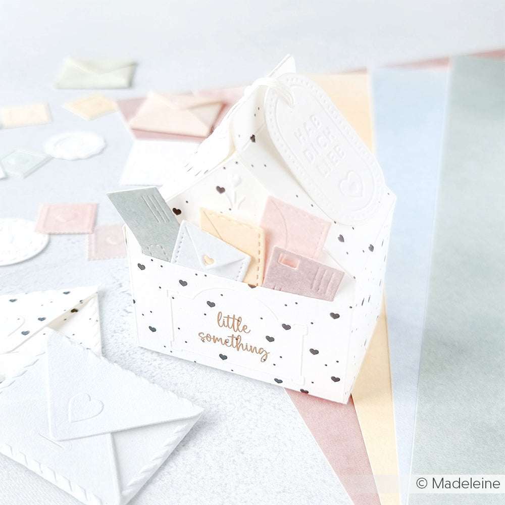Ein weißer Geschenkkarton aus Alexandra Renke-Papier mit einer "Kleinigkeit" enthält pastellfarbene Mini-Umschläge und -Karten, die mit einem Cut & Emboss Folder bearbeitet und auf einer hellen Oberfläche präsentiert werden.
