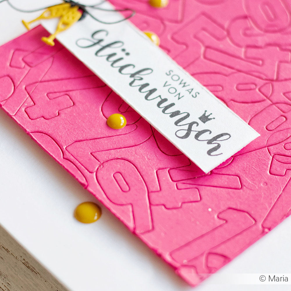 Nahaufnahme einer rosafarbenen Prägekarte aus 700g/m² Uni Cardstock von Alexandra Renke, mit Zahlen und einem weißen "Glückwunsch"-Schriftzug.