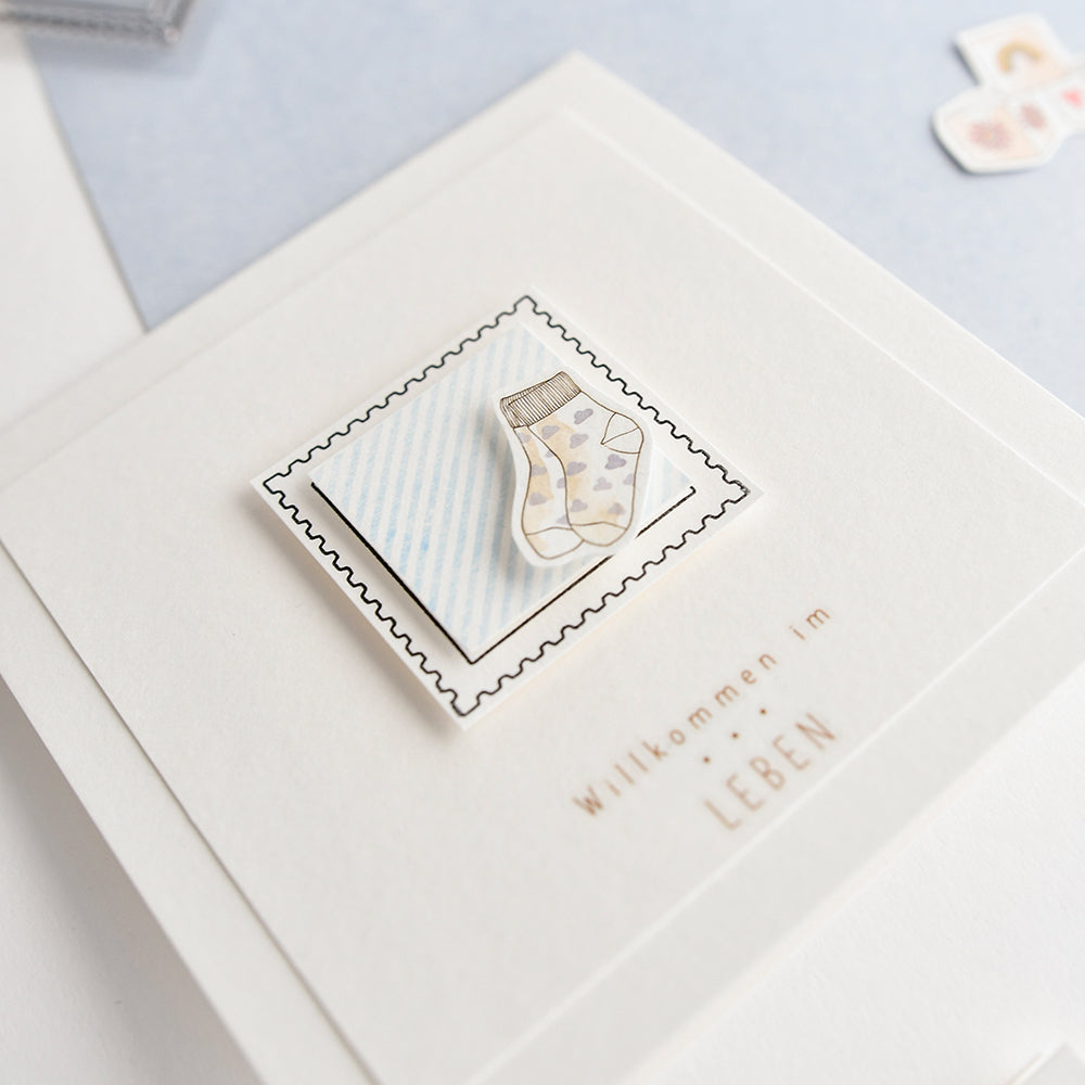 Eine Karte mit einer Zeichnung von Babysocken, die mit einem Photopolymer-Klarsichtstempel von Alexandra Renke hergestellt wurde, und dem deutschen Text "Willkommen im Leben".