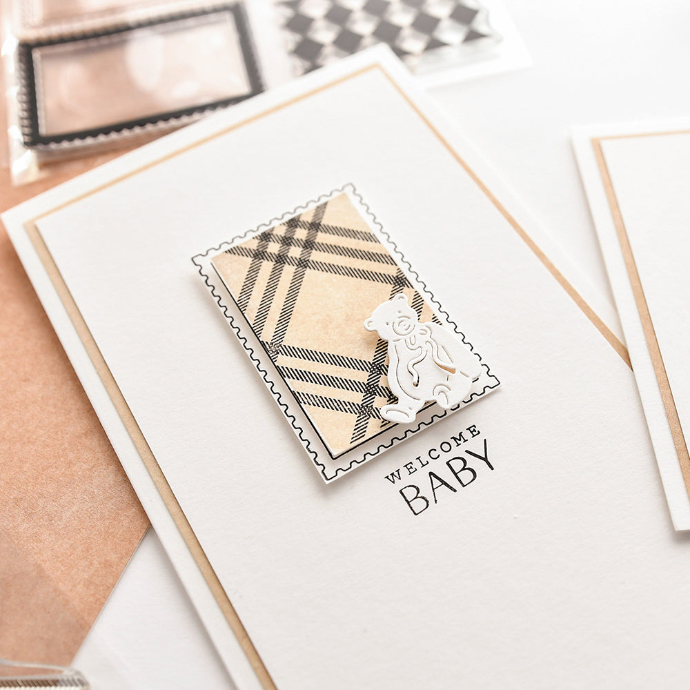 Grußkarte von Alexandra Renke mit kariertem Karomuster-Klarsichtstempel, Teddybär-Design und Text WELCOME BABY auf weißem Hintergrund.