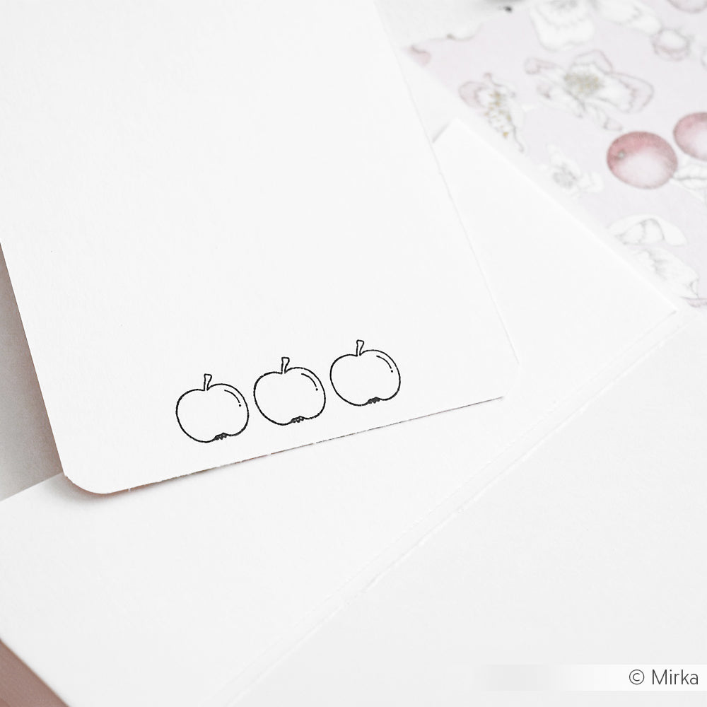 Eine weiße Karte von Alexandra Renke mit drei einfachen Zeichnungen von Äpfeln am unteren Rand, die mit einem Photopolymer-Stempel hergestellt wurden, ruht auf anderen Papieren.