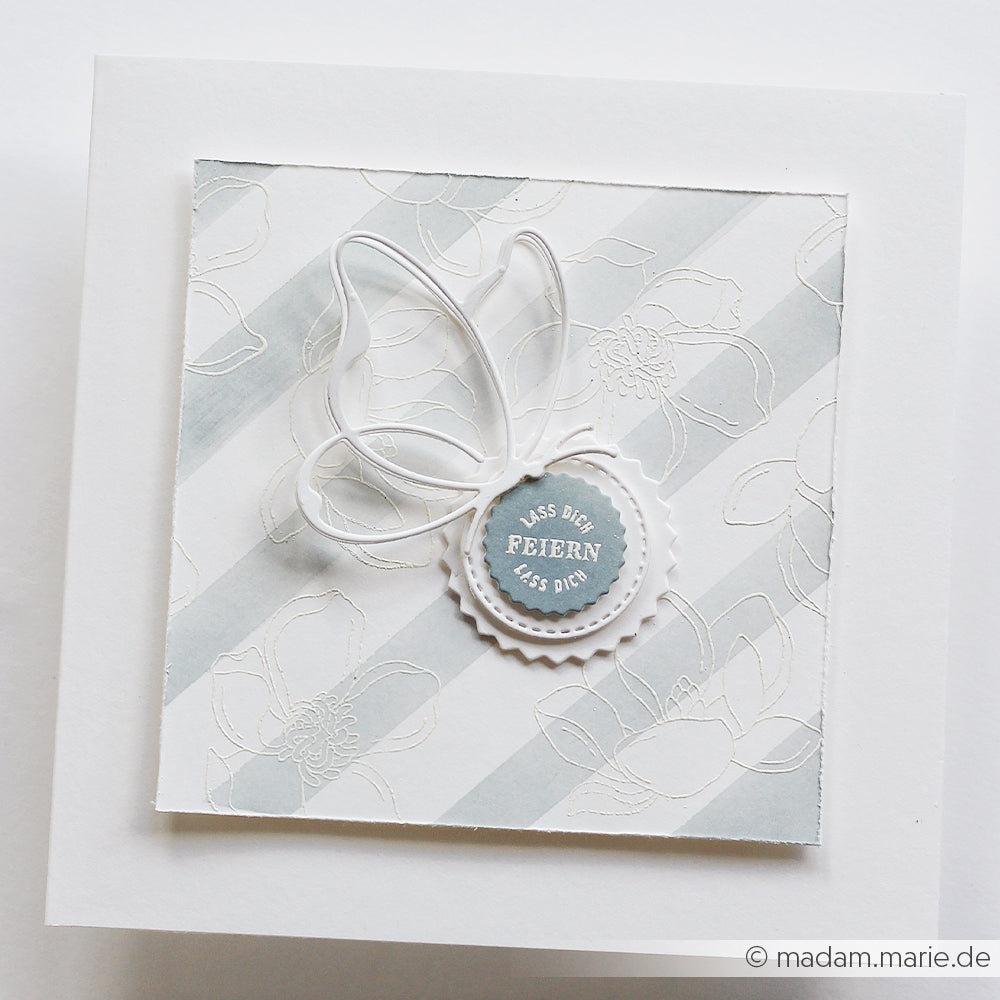 Quadratische Karte von Alexandra Renke mit weißem Blumenmuster, diagonalen Streifen, Schleife und rundem "Lass Dich feiern"-Label. Veredelt mit Sommerblüten-Motiven in klarer Photopolymer-Stempeltechnik für zusätzliche Eleganz.