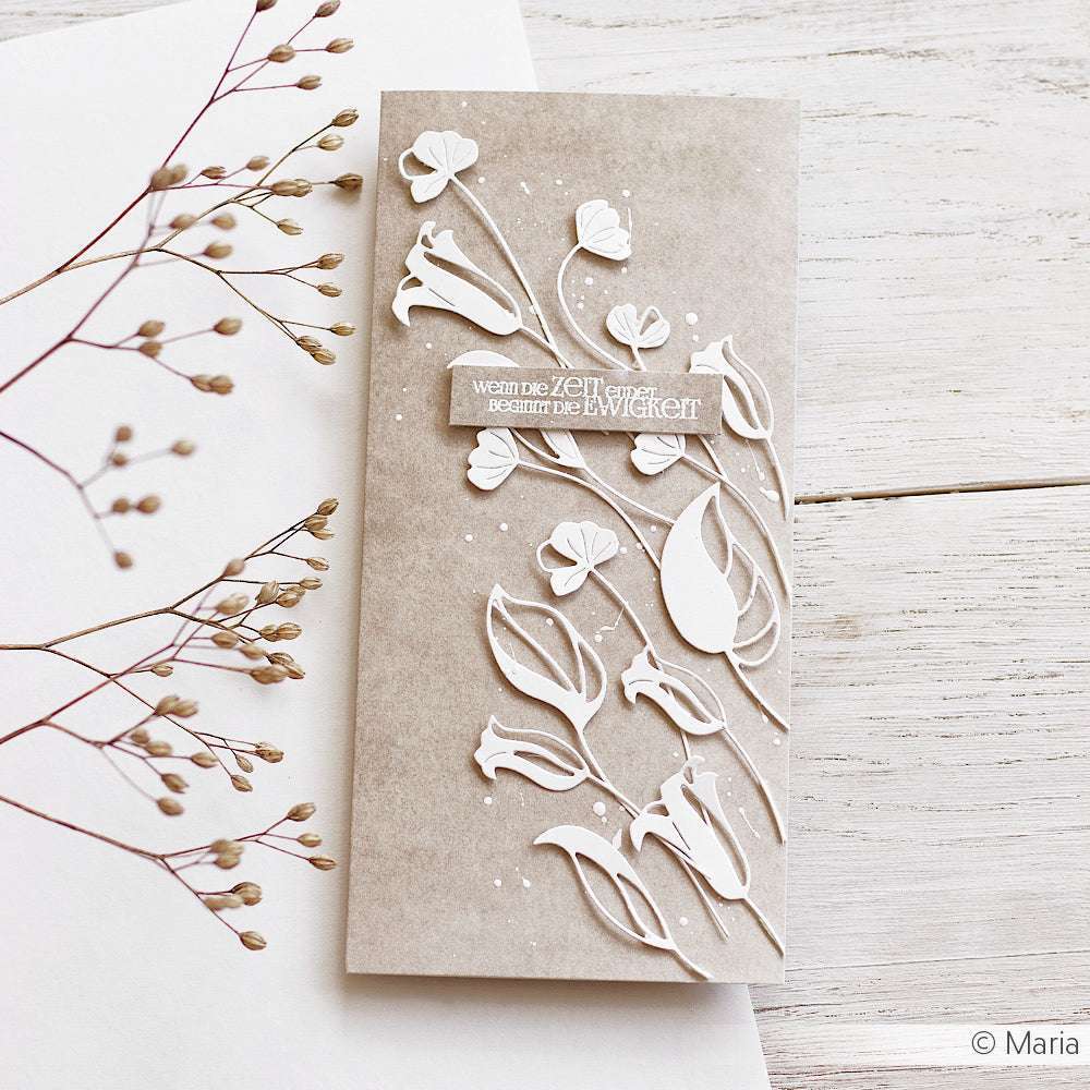 Handgefertigte beige Karte mit weiß geprägten Blumenmotiven, hergestellt mit Alexandra Renke Designpapieren und einer Motivstanze Fransenblume, neben zarten getrockneten Pflanzenstängeln auf weißem Untergrund.