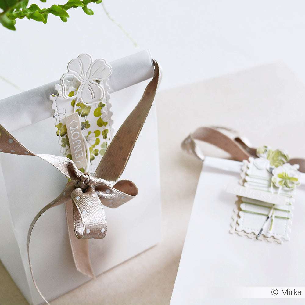 Alexandra Renke weiße Geschenktüten mit beigen Bändern, dekorativen Anhängern und Fotopolymer-Stempelakzenten, die auf einer hellen Oberfläche für einen weichen, eleganten Look sorgen.