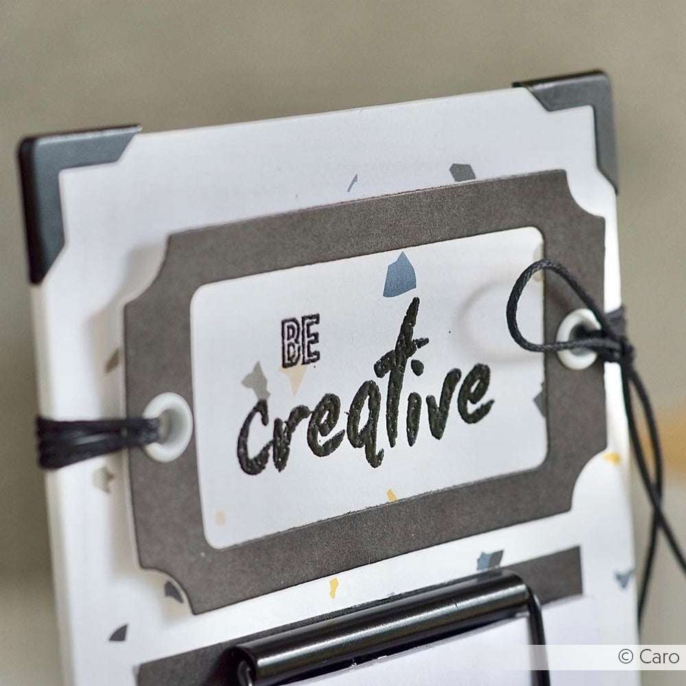 Ein Klemmbrett mit einer "Be creative"-Karte in fetten, künstlerischen Buchstaben, die von einer schwarzen Schnur gehalten wird - inspirierend für Ihr nächstes Alexandra Renke Clear Stamp Set oder Photopolymer Stempel Projekt.