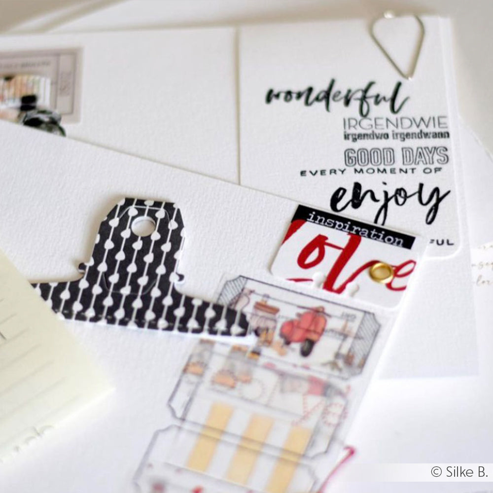 Ein Schreibtisch mit Scrapbook-Papier von Alexandra Renke, dekorativen Anhängern, Textaufklebern, einer schwarzen gemusterten Papierklammer und einem Satz klarer Stempel - perfekt, um Ihre Projekte mit individuellen Designs zu versehen.