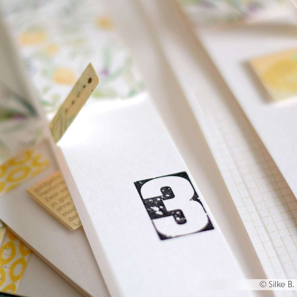 Weißes Papier mit einer auffälligen schwarzen Zahl 3, umgeben von gemustertem Papier und Registerkarten - die perfekte Inspiration für die Verwendung von Gebrochenen Zahlen oder einem Alexandra Renke Photopolymer Stempel in Ihren kreativen Projekten.
