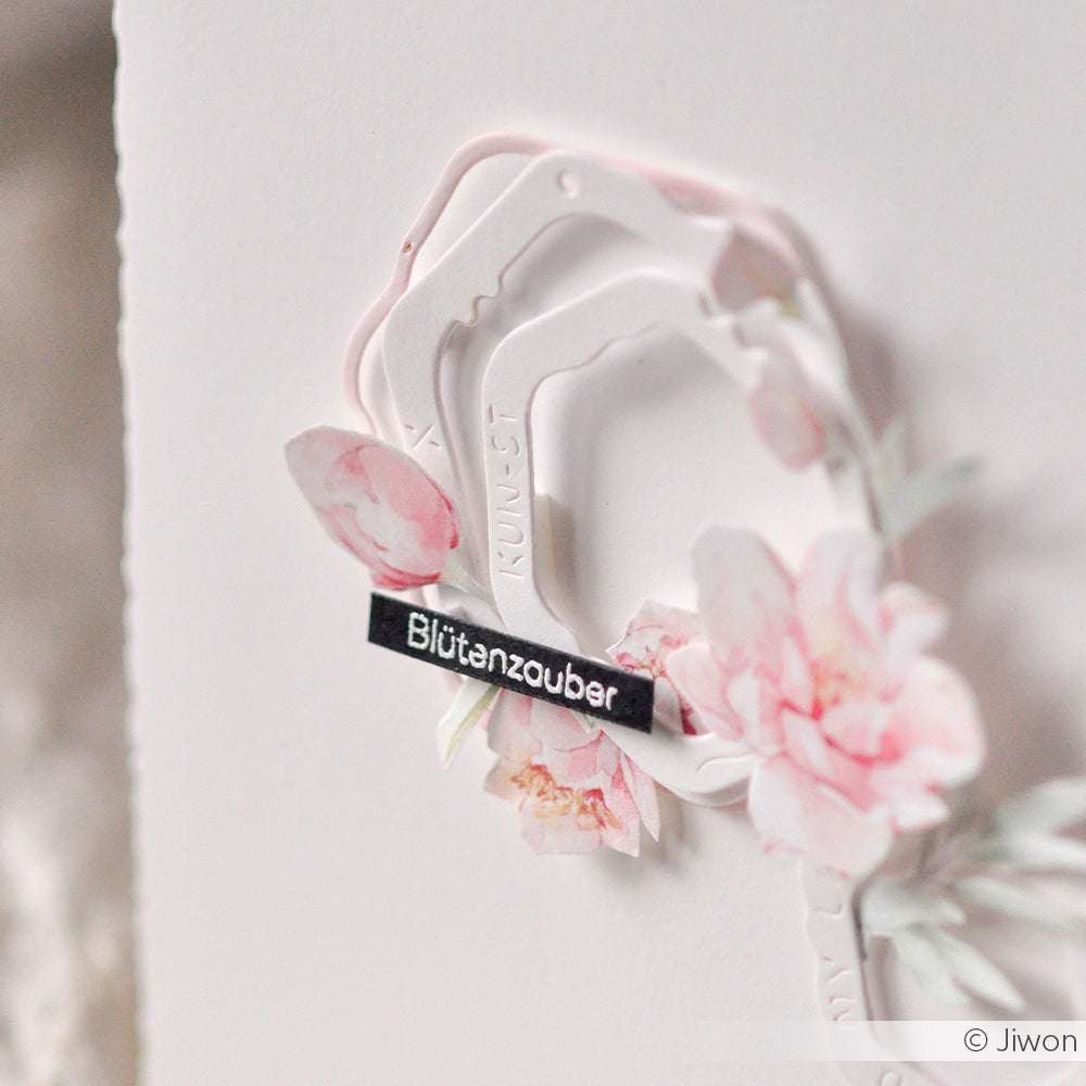 Weiße Karte mit geschichteten Papierrahmen, rosafarbenen Aquarellblumen und einem schwarzen "Blütenzauber"-Etikett, das mit Alexandra Renke Photopolymer Stempel für scharfe und detaillierte Blumenakzente erstellt wurde.