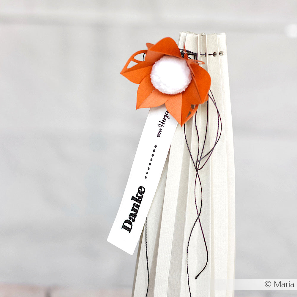 Orangefarbene Papierblume mit weißem "Danke"-Anhänger, gestempelt mit dem Clear Stamp Danke von Alexandra Renke, befestigt an einem gefalteten weißen Fächer, der mit dunklen Schnüren gebunden ist.
