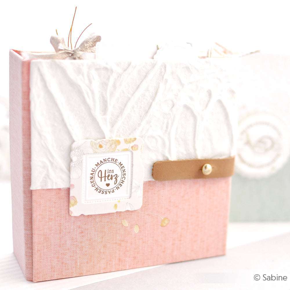 Eine rosa-weiße, handgefertigte Geschenkbox mit strukturiertem Papier, einem kleinen Anhänger, einem Lederband und einem klaren Alexandra Renke-Stempel für eine persönliche Note.