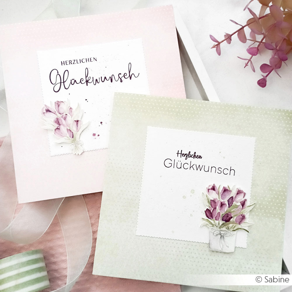 Zwei pastellfarbene Grußkarten von Alexandra Renke mit floralen Verzierungen und dem deutschen Text "Blumen für dich", die mit klaren Stempeln hergestellt wurden, werden auf einer hellen Fläche mit einem Band in der Nähe ausgestellt.
