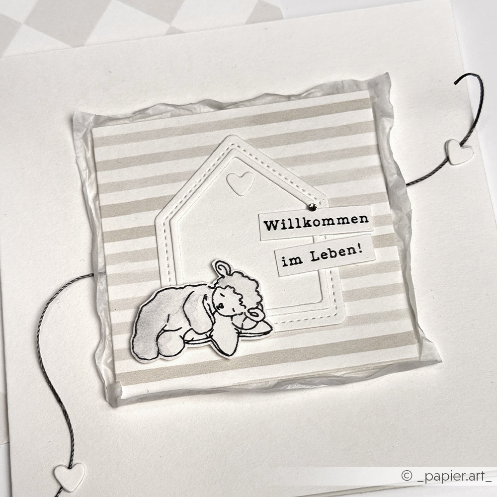 Grußkarte von Alexandra Renke mit einem Teddybär, einem Hausumriss und dem Text "Willkommen im Leben!" auf einem grau gestreiften Hintergrund, erstellt mit einem hochdetaillierten Photopolymerstempel.