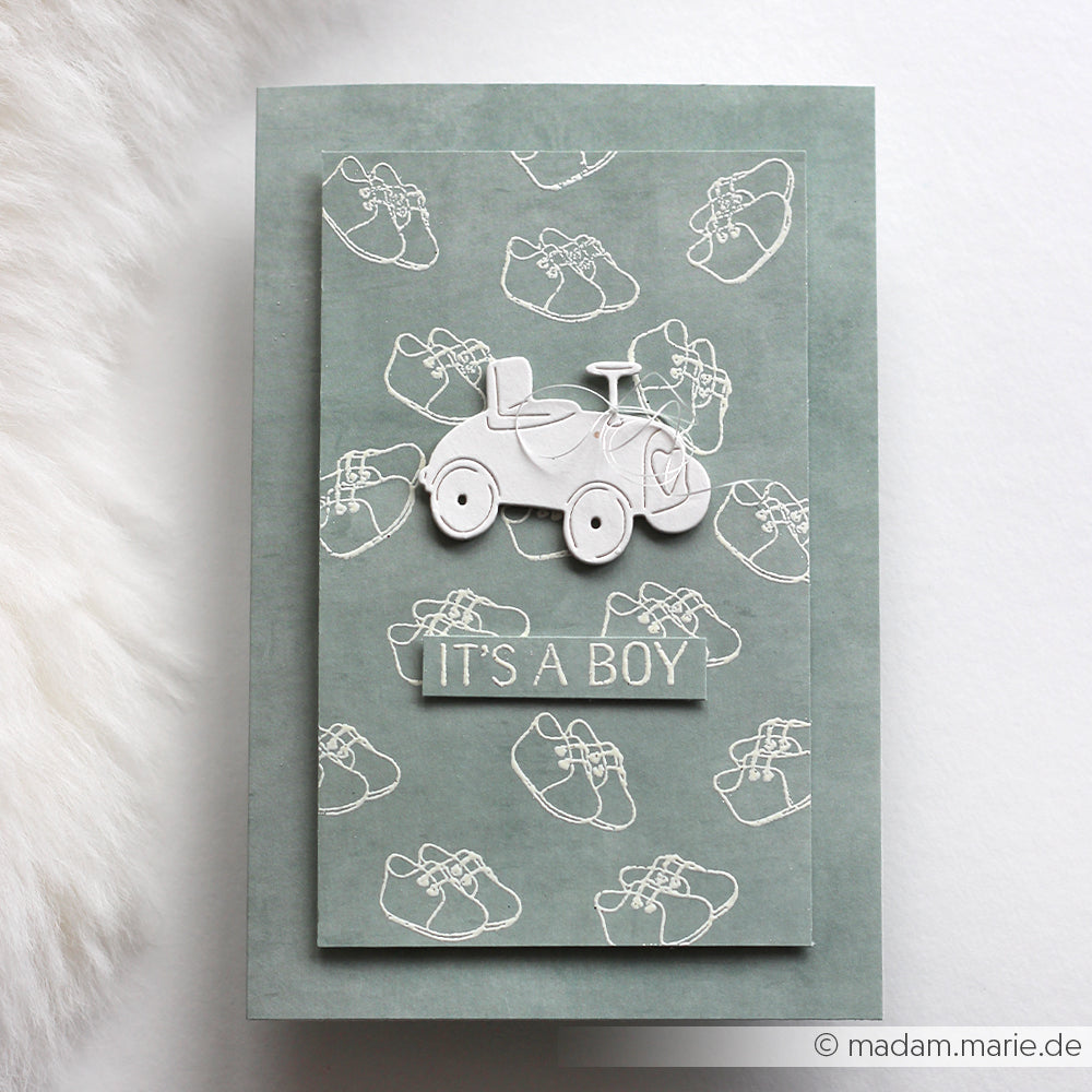 Die grüne Karte von Alexandra Renke zeigt weiße Umrisse von Babyschuhen, eine Illustration eines Spielzeugautos und den Text "It's a boy" - ideal, um sie mit Ihren Lieblingsmotiven von Clear Stamp oder Photopolymer Stempel zu kombinieren.