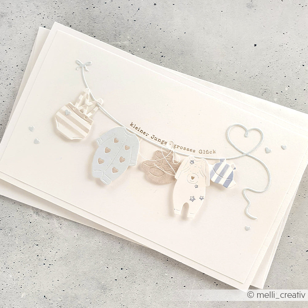 Handgefertigte Karte von Alexandra Renke mit winzigen Babykleidern und Herzen an einer Schnur, mit dem deutschen Text "Kleines Junge, großes Glück" und einem klaren "It's a boy"-Stempel für eine besondere Note.