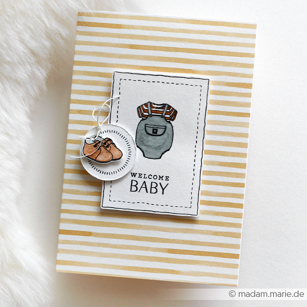 Grußkarte von Alexandra Renke mit beigefarbenen Streifen, Illustration von Babykleidung und dem Text "Komposition Welcome Baby". Ideal zum Kombinieren mit Ihren Lieblingsmotiven aus photopolymeren Stempeln oder Klarsichtstempeln.