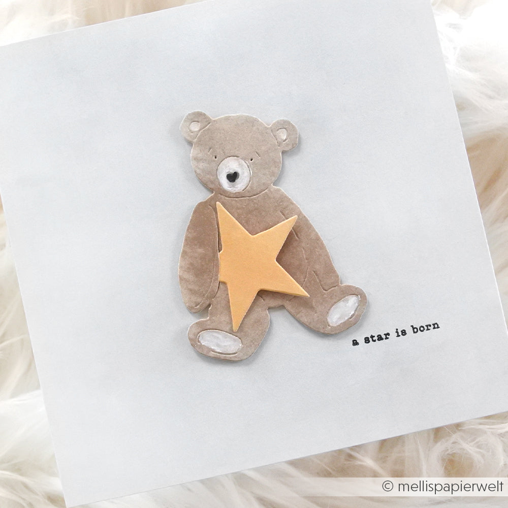 Eine handgefertigte Karte von Alexandra Renke zeigt einen braunen Teddybären mit einem gelben Stern und dem Text "a star is born", der mit Clear Stamp für gestochen scharfe Details hergestellt wurde - ein ideales Willkommens-Baby-Andenken.