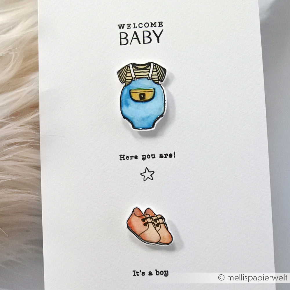 Eine Karte aus der Komposition Welcome Baby von Alexandra Renke zeigt einen blauen Overall, braune Schuhe und einen Clear Stamp mit dem Text: Hier bist du! Es ist ein Junge.