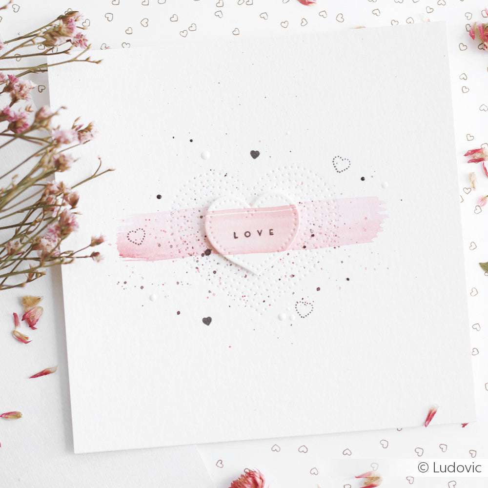 Quadratische Karte von Alexandra Renke mit einem rosafarbenen Herz mit der Aufschrift LOVE, das mit kleinen Herzen und Trockenblumen verziert ist. Die Karte ist aus Designpapier gefertigt und mit zarten Stanzdetails versehen.