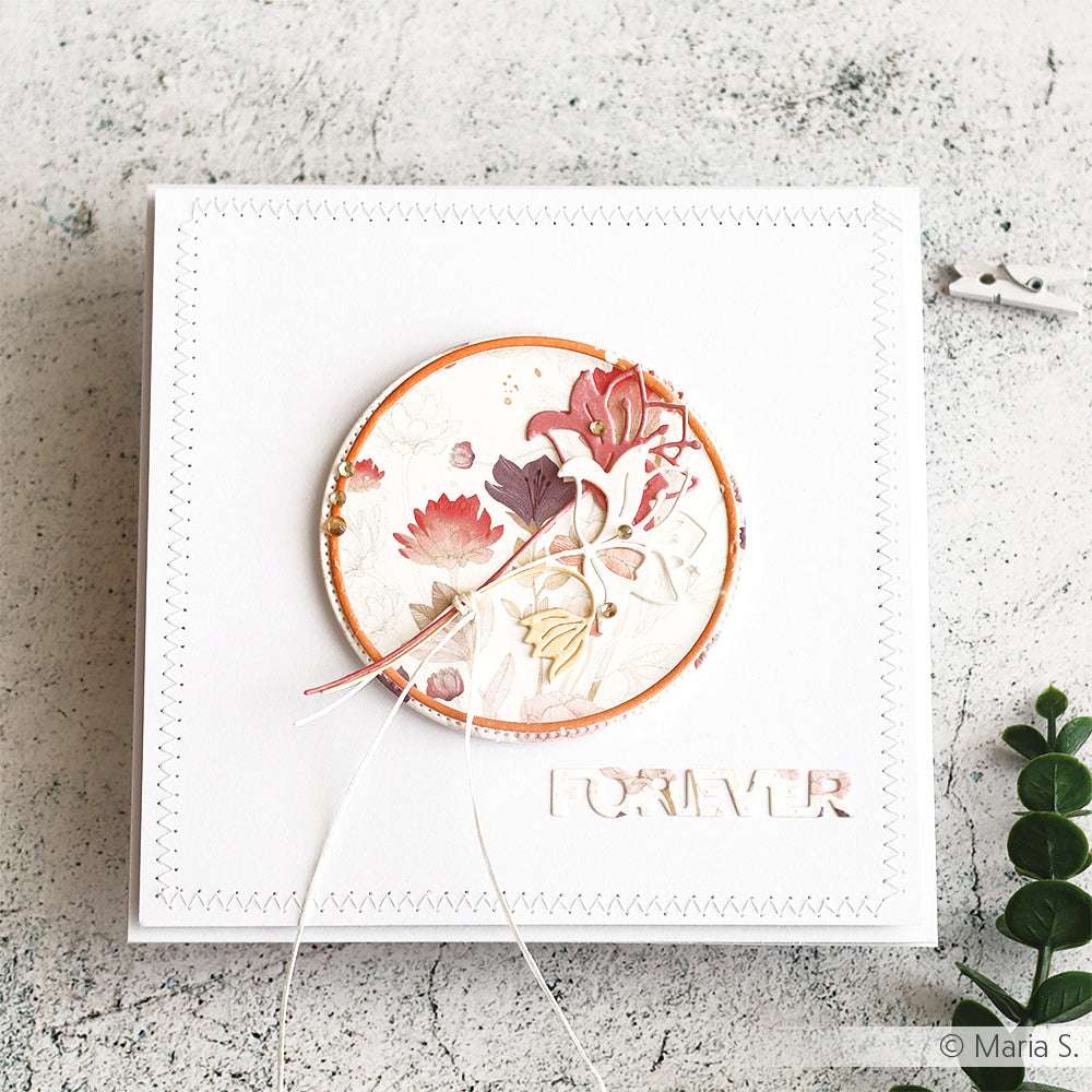 Die quadratische weiße Karte von Alexandra Renke zeigt ein florales Kreismuster und den Text FOREVER, der mit DieCut Systemen hergestellt wurde. Sie ruht auf einer strukturierten Oberfläche mit einem grünen Zweig in der Nähe.