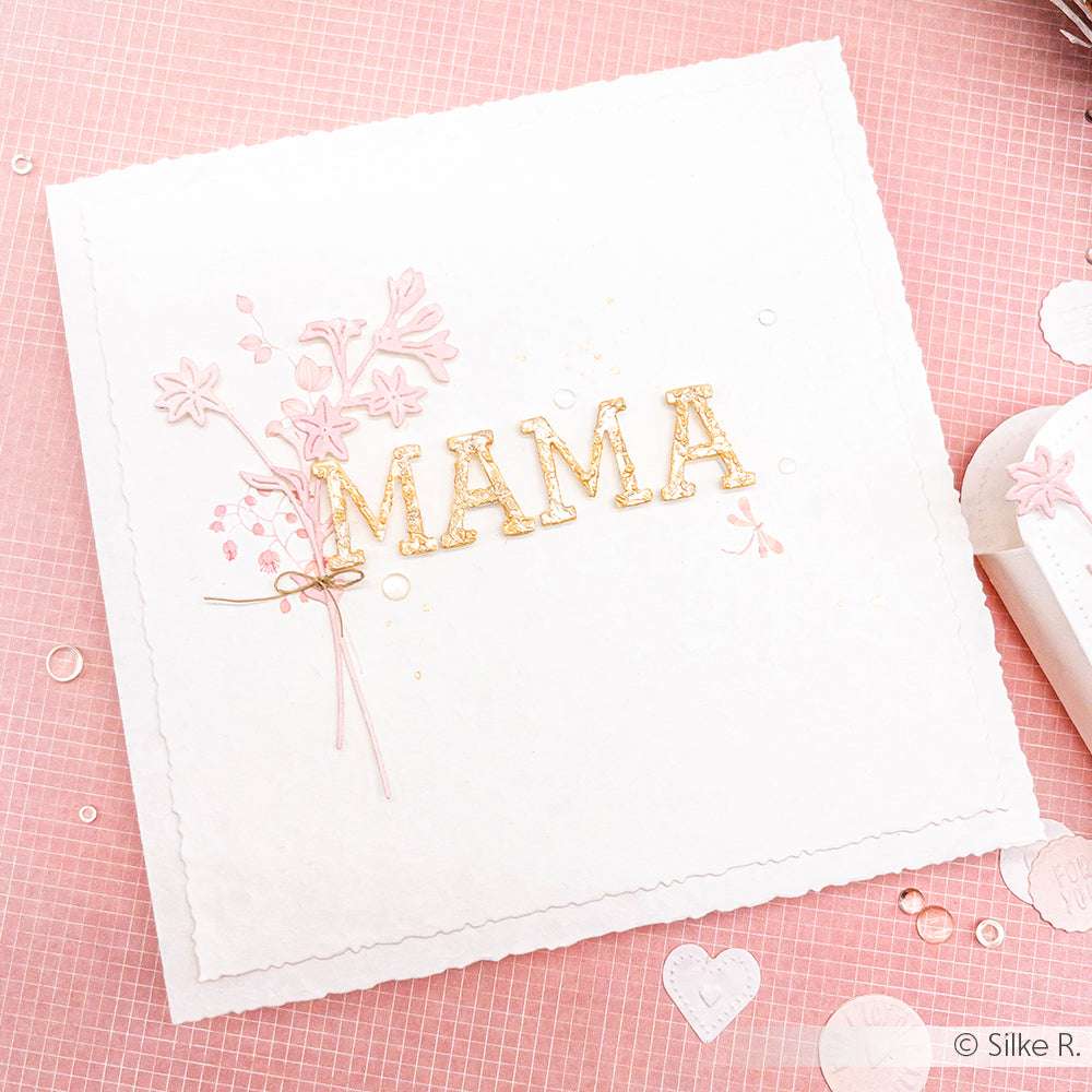 Quadratische weiße Karte mit "MAMA" in goldenen Buchstaben, hergestellt mit der Alexandra Renke Slab Alphabet Stanze, verziert mit rosa Blumen und einer Schleife, vor einem rosa strukturierten Hintergrund.