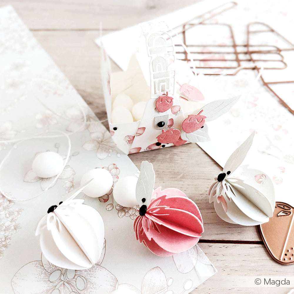 Bienenbasteleien aus Papier und eine dekorierte Schachtel mit weißen Bonbons liegen auf einem hellen Holztisch, auf dem im Hintergrund florale Designpapiere von Alexandra Renke zu sehen sind.