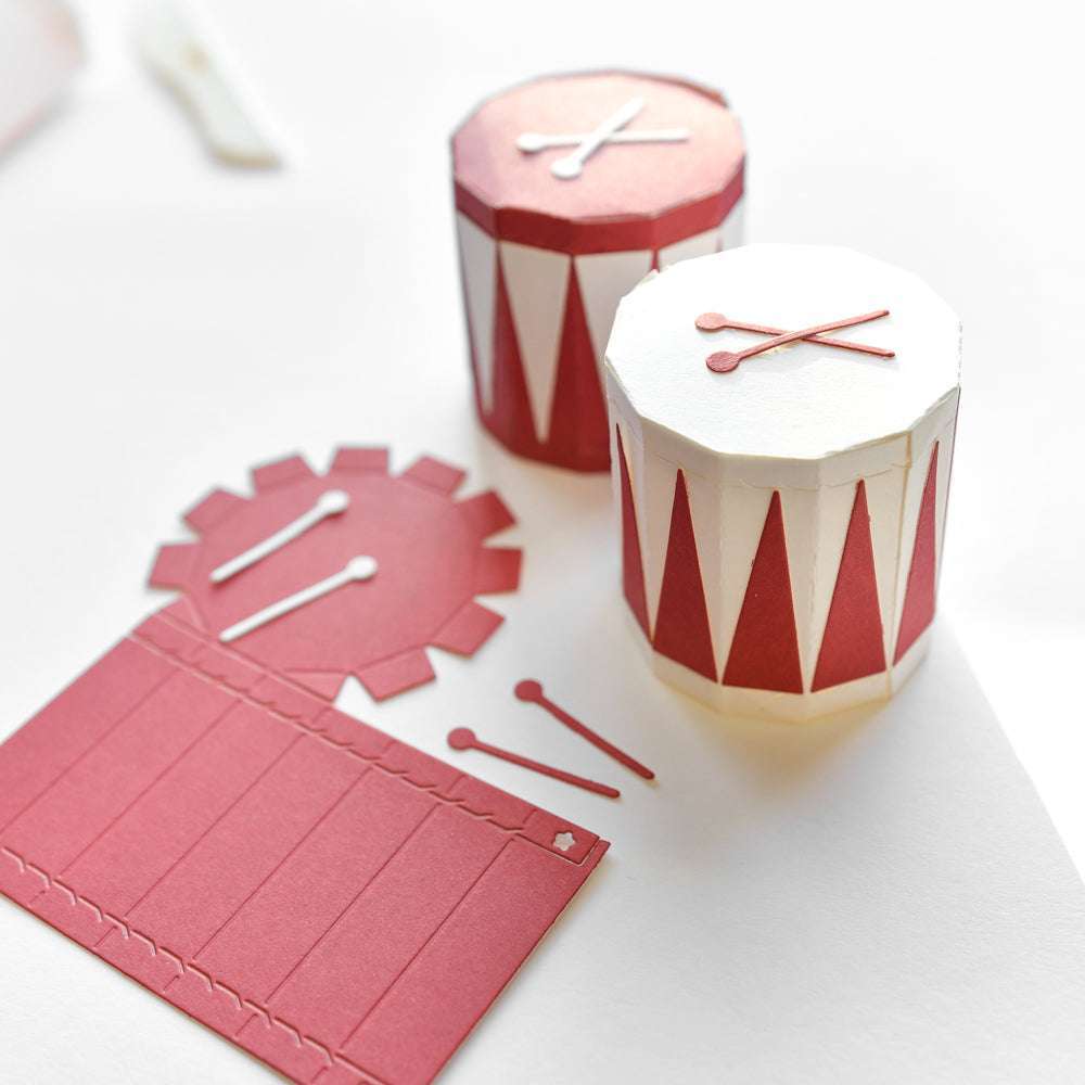 Zwei kleine, rot-weiße Papiertrommeln aus Alexandra Renke Designpapieren mit passenden Trommelstöcken ruhen auf einer weißen Fläche - ideal für Ihr nächstes Projekt mit der Motivstanze Trommelbox.